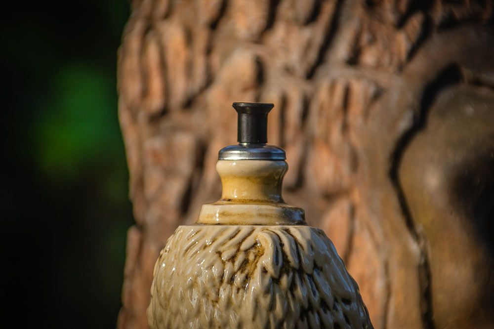 Primer plano de un objeto de cerámica texturizada de color claro con un pequeño tapón cilíndrico negro, sobre un fondo de árbol borroso.