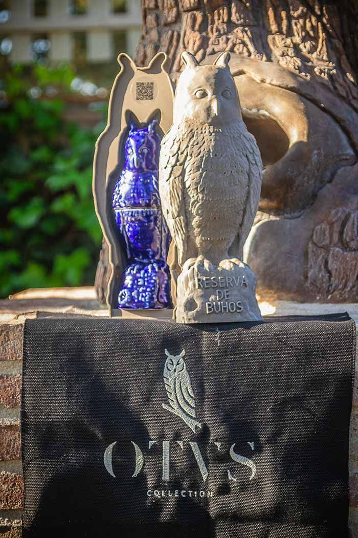 Una escultura de piedra gris con forma de búho junto a un búho azul iridiscente dentro de una caja con la etiqueta