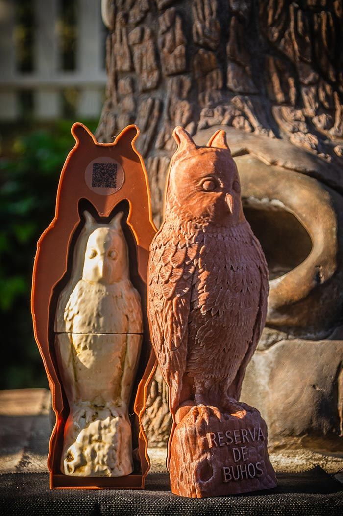 Un molde de silicona marrón con forma de búho se encuentra junto a una escultura de arcilla terminada con forma de búho, sobre una superficie texturizada que imita la corteza de un árbol.