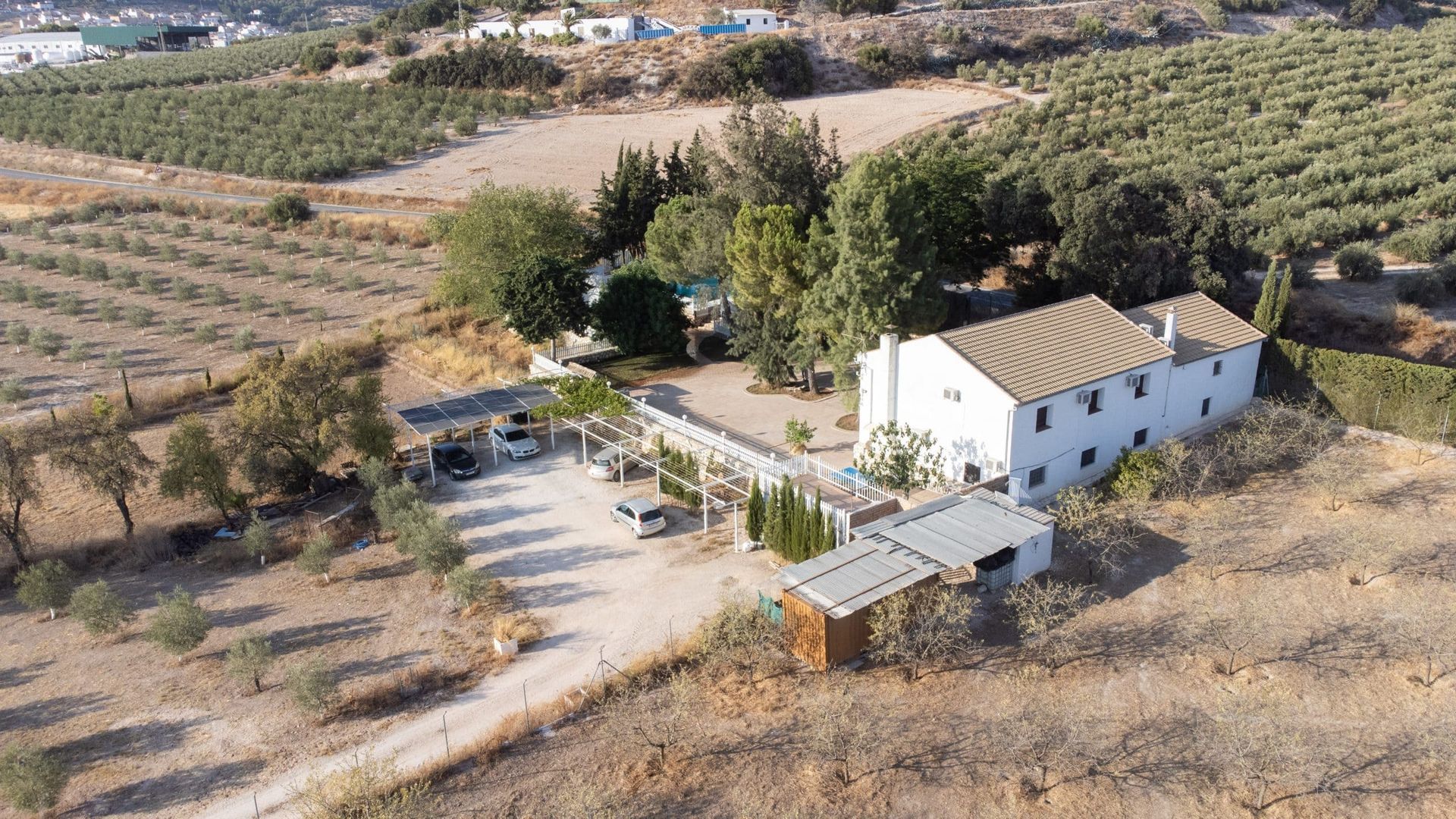Vista aérea de un edificio blanco con cocheras, autos estacionados, árboles y un camino de tierra en un entorno rural seco.