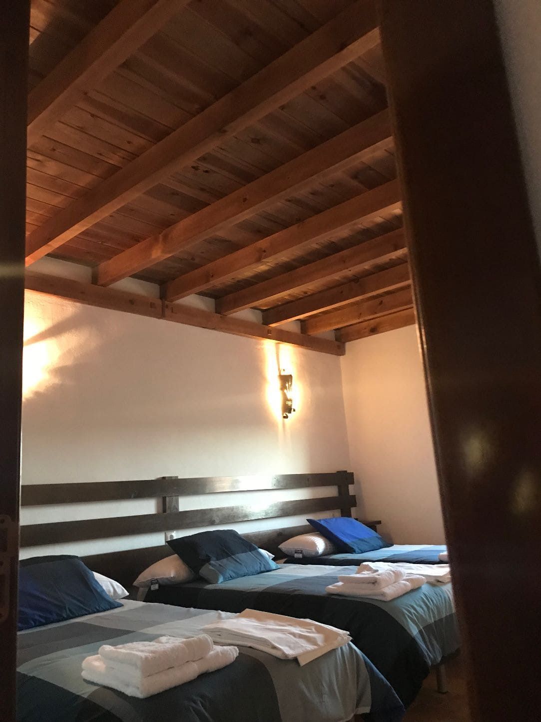 Dormitorio con tres camas; techo de madera, paredes blancas, ropa de cama azul y toallas blancas.