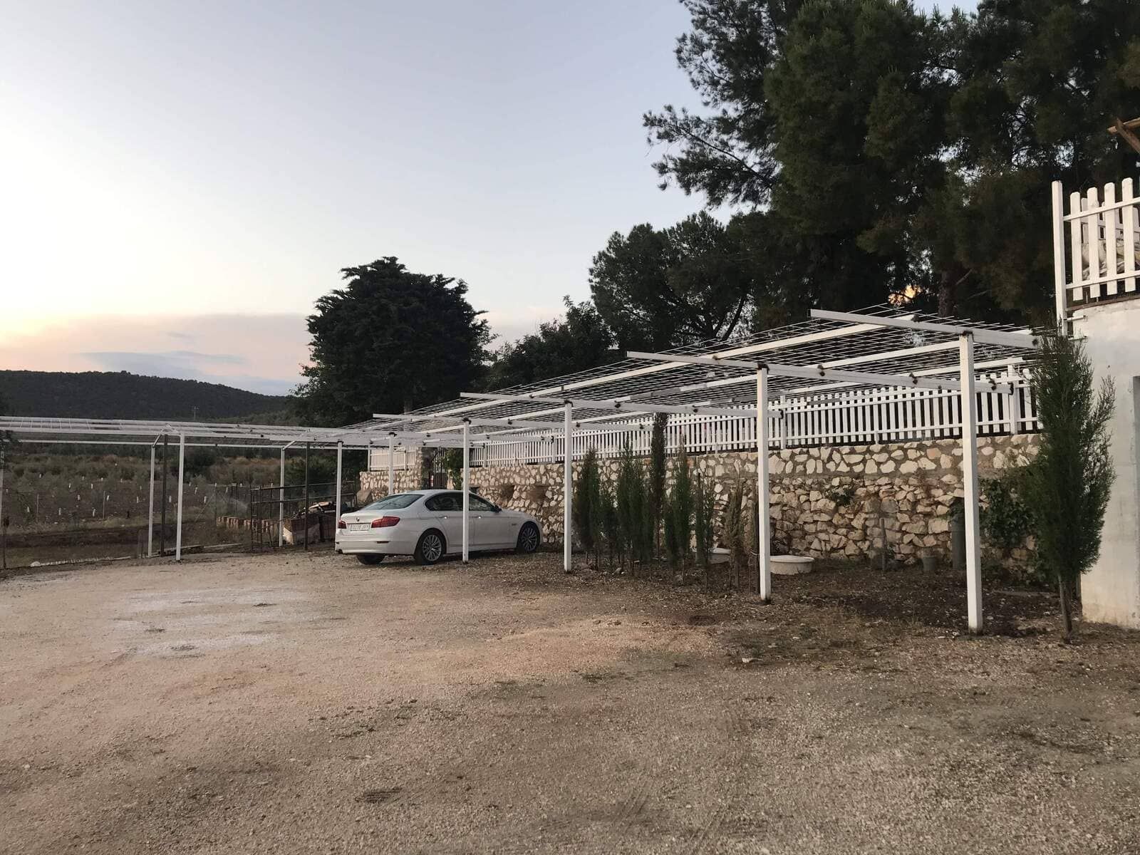 Coche aparcado bajo una pérgola metálica blanca, junto a un muro de piedra y una valla, en un entorno exterior al anochecer.