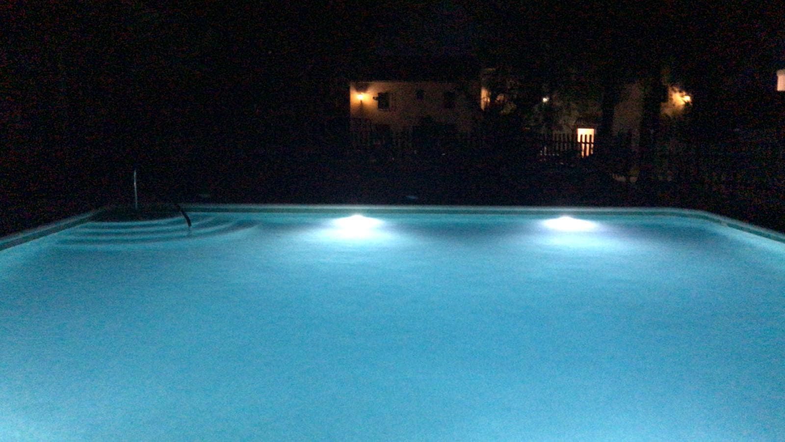 Piscina iluminada de noche con tres luces subacuáticas. Se ven edificios en el fondo oscuro.