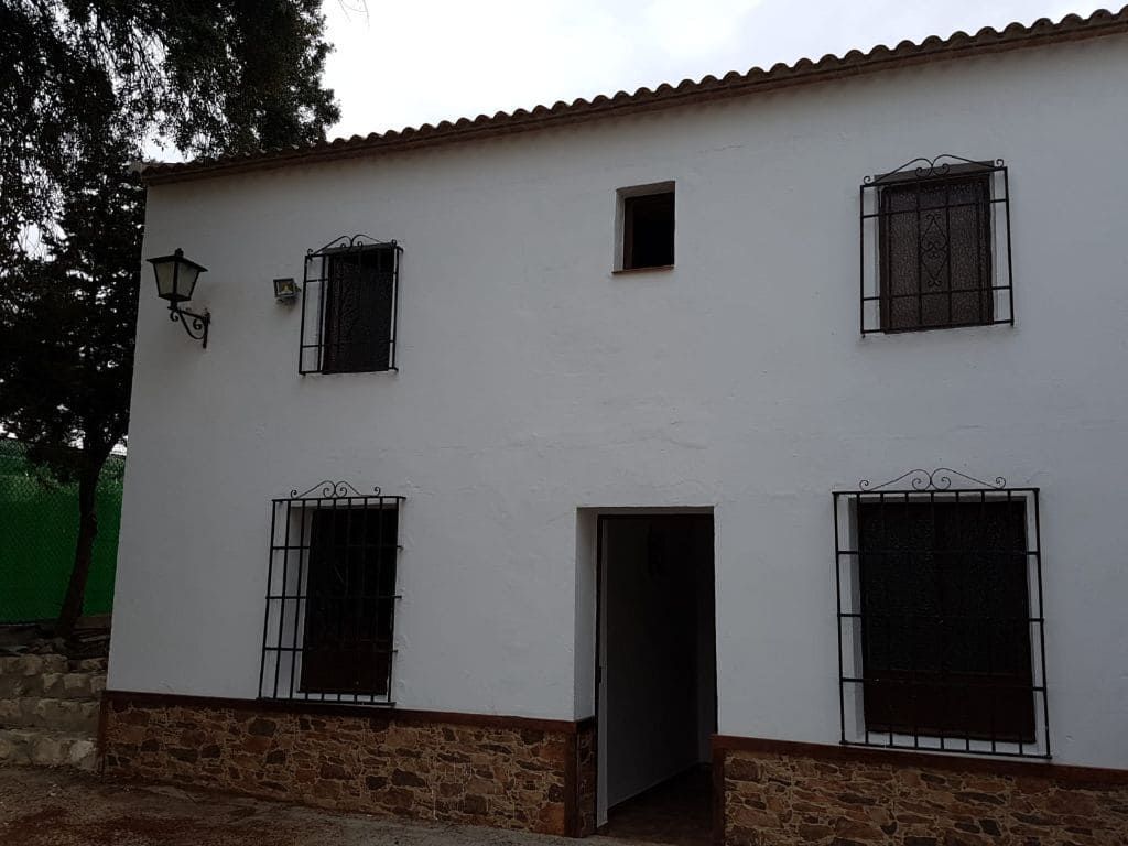 Edificio blanco de dos pisos con ventanas enrejadas, base de ladrillo y puerta abierta bajo un cielo nublado.