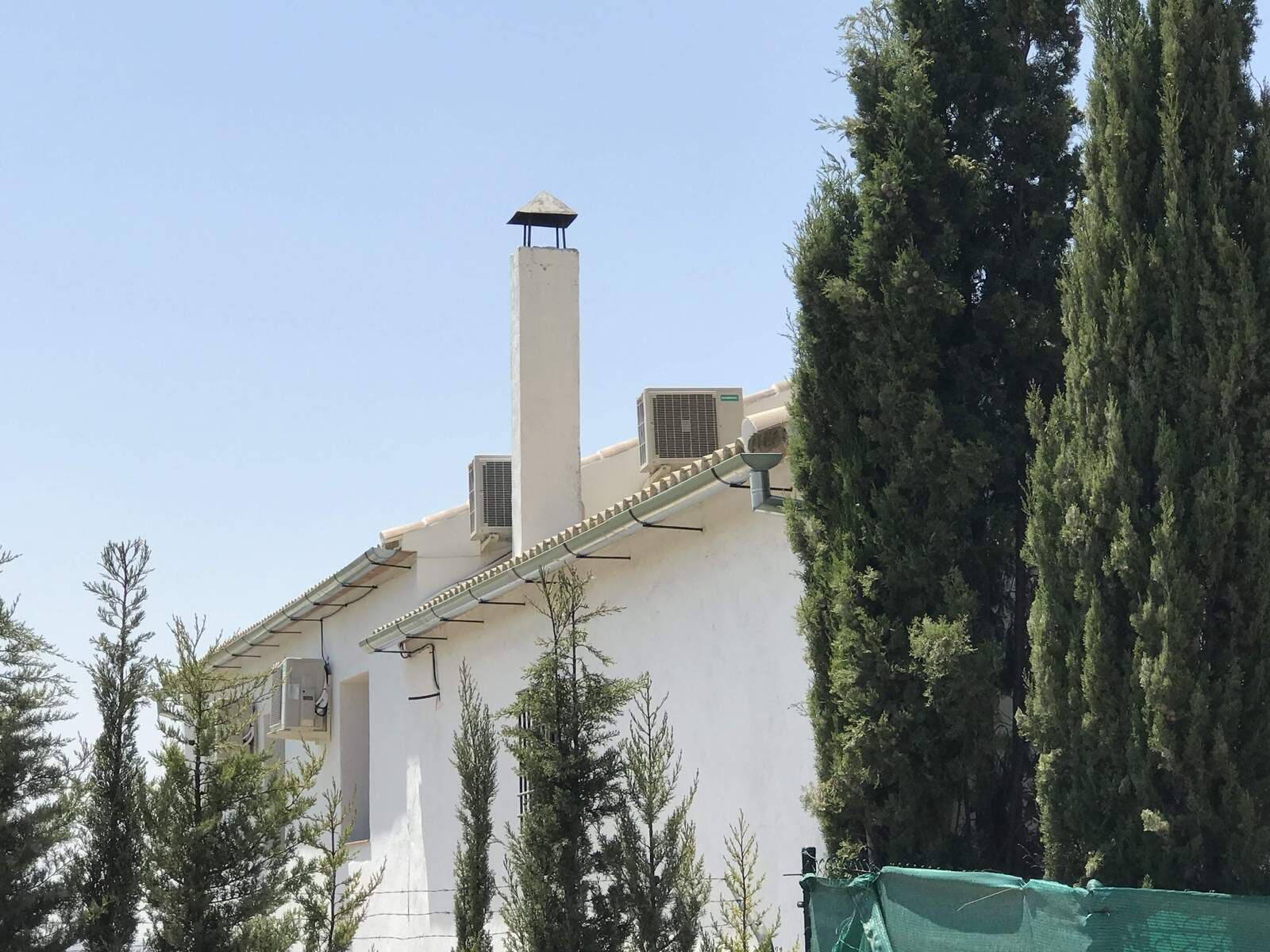 Edificio blanco con chimenea, aire acondicionado y altos árboles verdes contra un cielo azul.