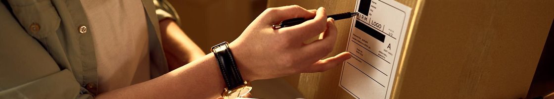 Una persona con un reloj escribe en una caja de cartón con un bolígrafo negro.