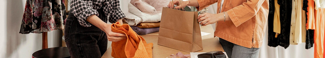 Dos personas en una tienda de ropa, una de ellas metiendo una prenda naranja en una bolsa de papel marrón. Percheros al fondo.