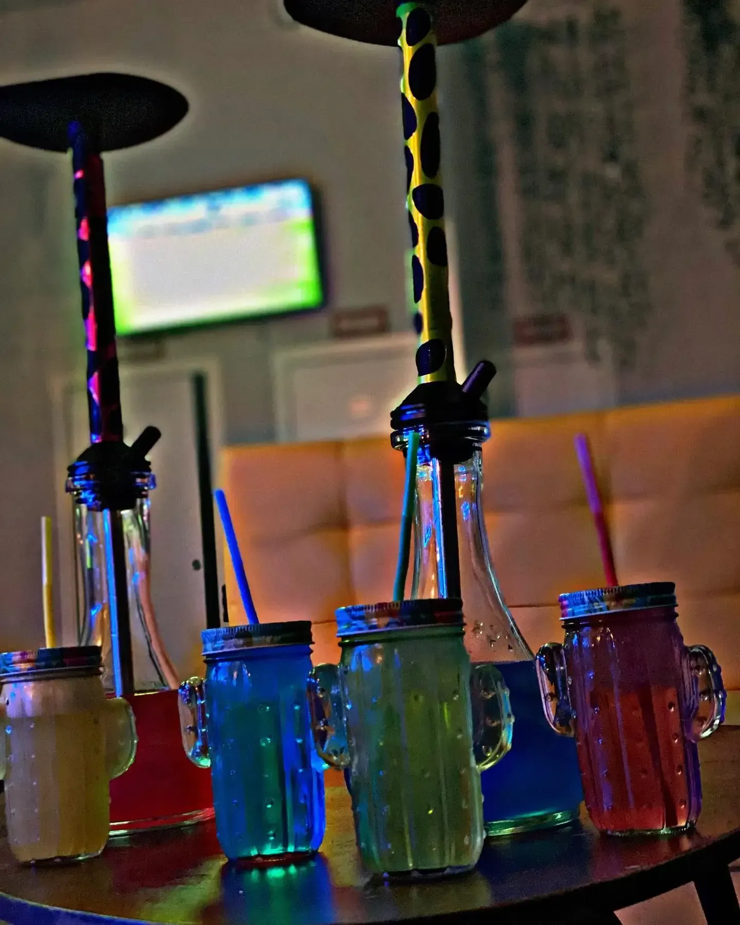 Narguiles y bebidas en vasos con forma de cactus sobre una mesa. Colores neón iluminan la escena.