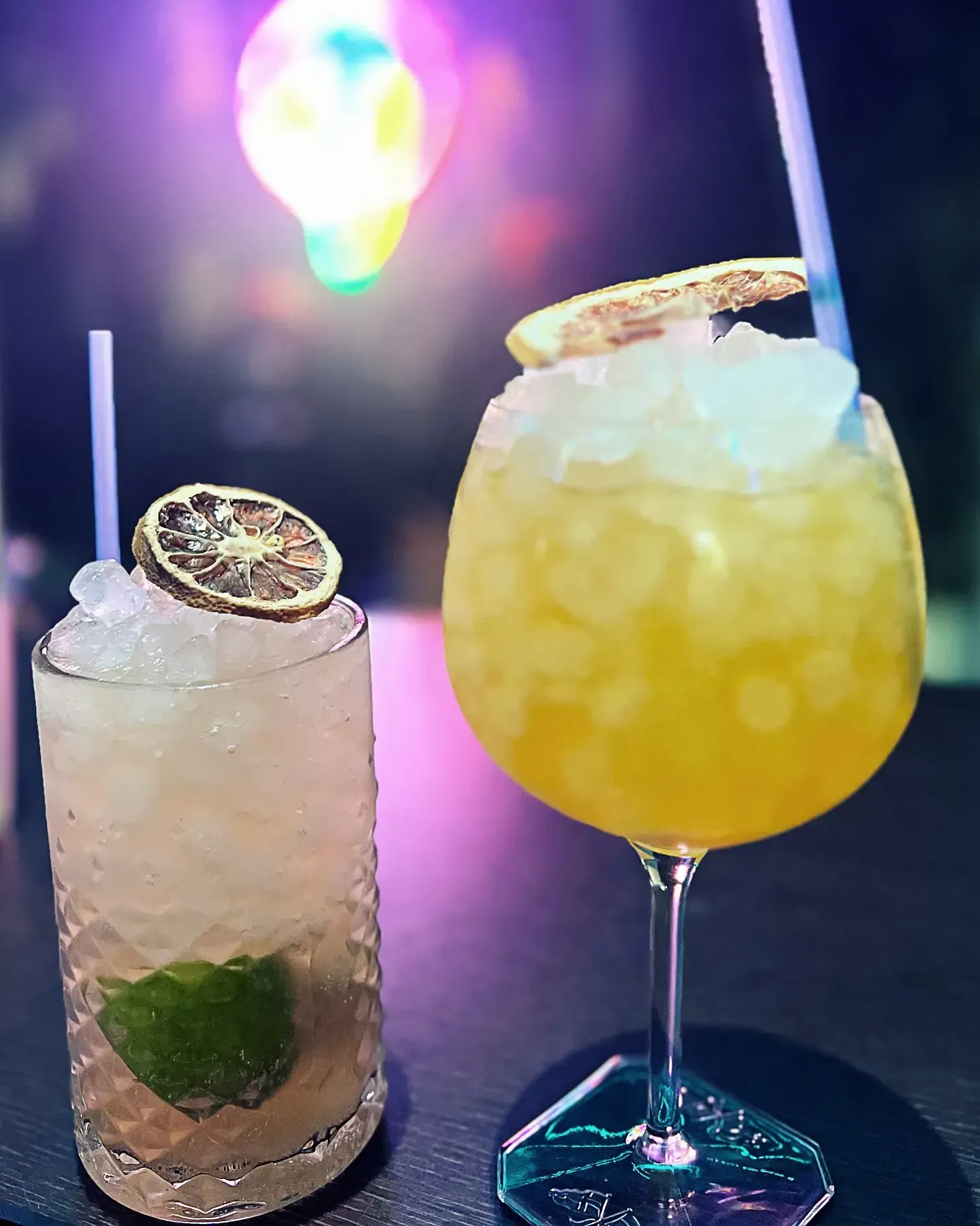 Dos cócteles, uno transparente y otro amarillo, con hielo y guarniciones, en una barra.