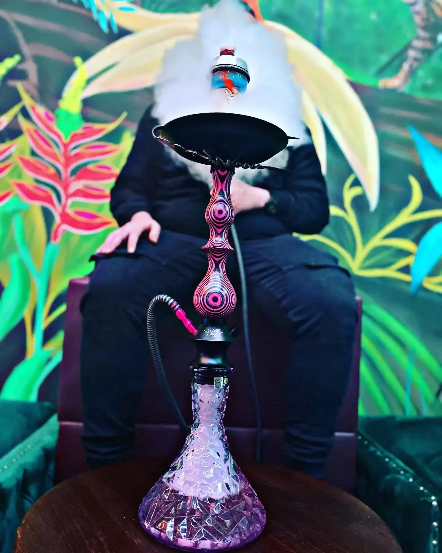Persona oculta por un objeto blanco, sentada cerca de una pipa de agua morada, sobre un fondo de mural floral.
