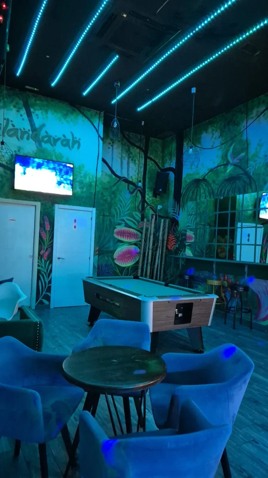 Mesa de billar y asientos en un bar iluminado de azul con un mural de temática jungla y luces de neón.