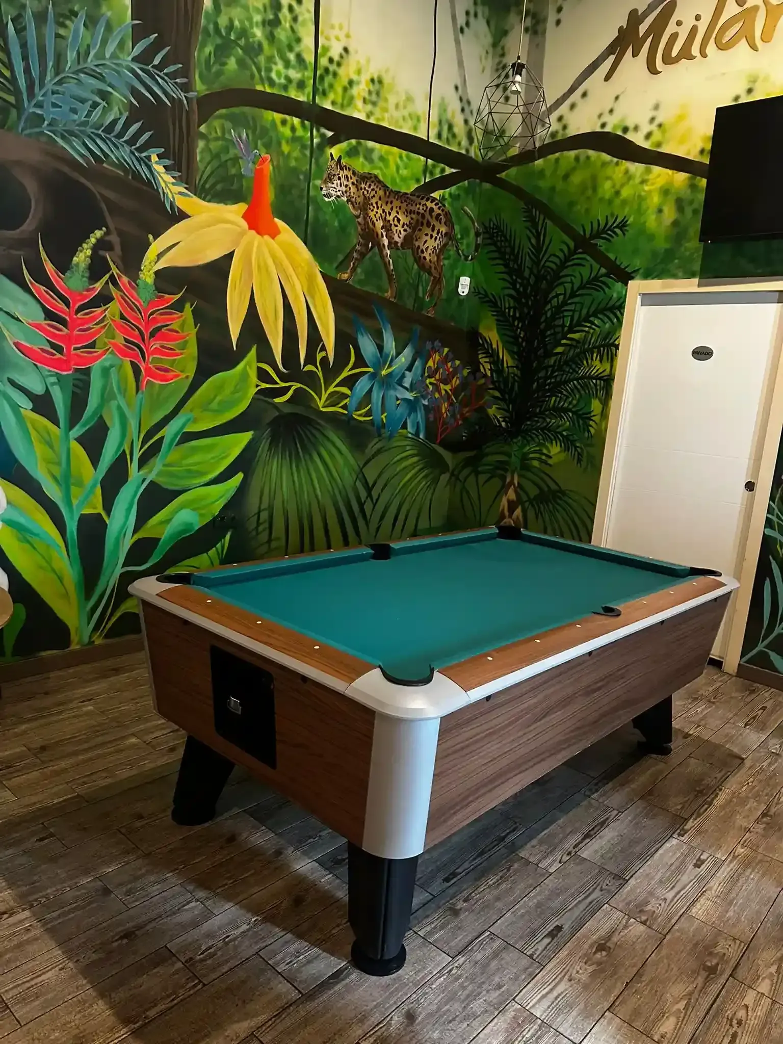Mesa de billar con fieltro verde y detalles de madera marrón, sobre un mural con temática de jungla.