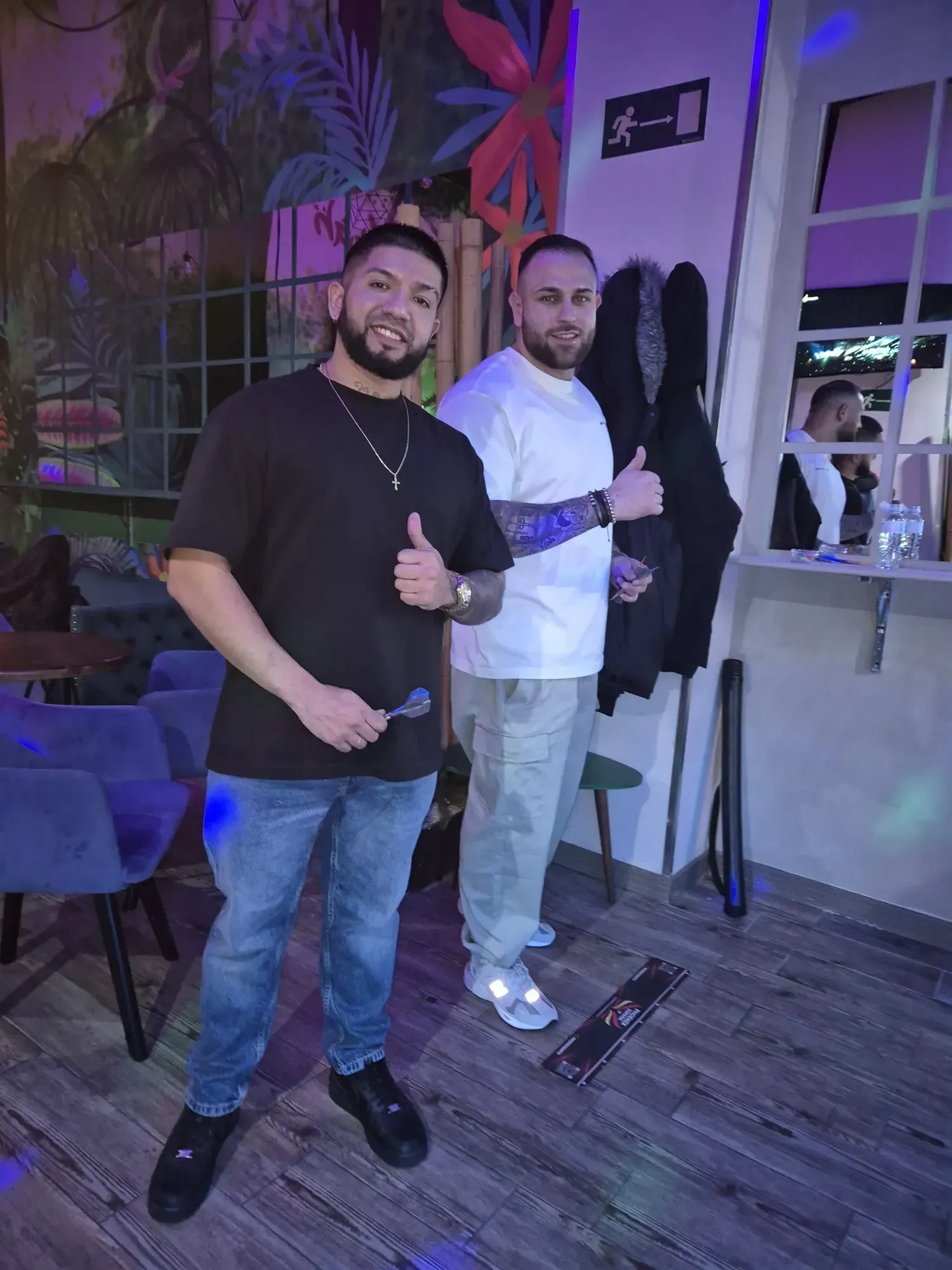 Dos hombres levantan el pulgar en un bar con un mural tropical. Uno viste de negro, el otro de blanco.