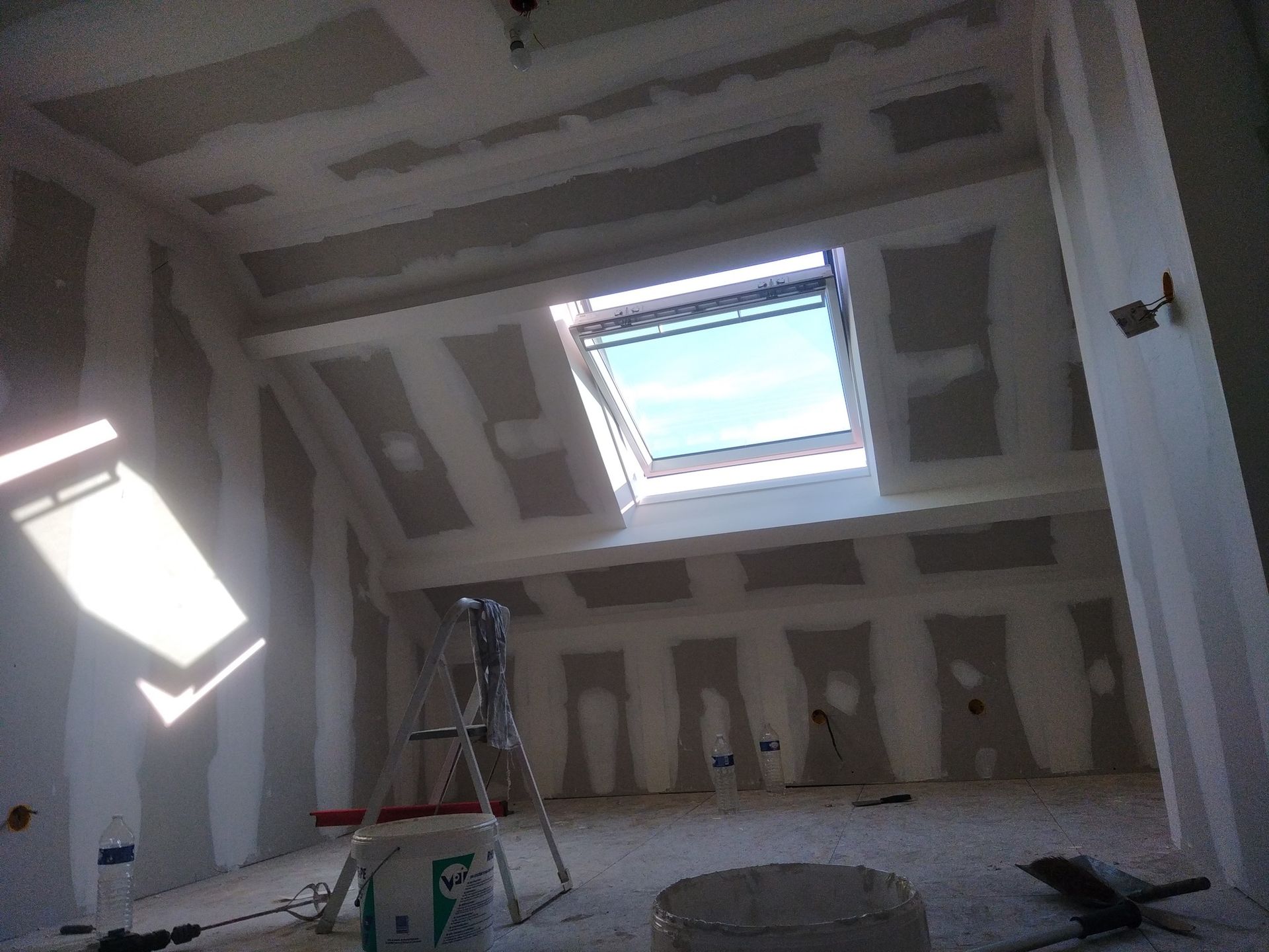 Intérieur d'une pièce inachevée avec des cloisons sèches, un puits de lumière, une échelle, un seau et de la lumière qui entre.