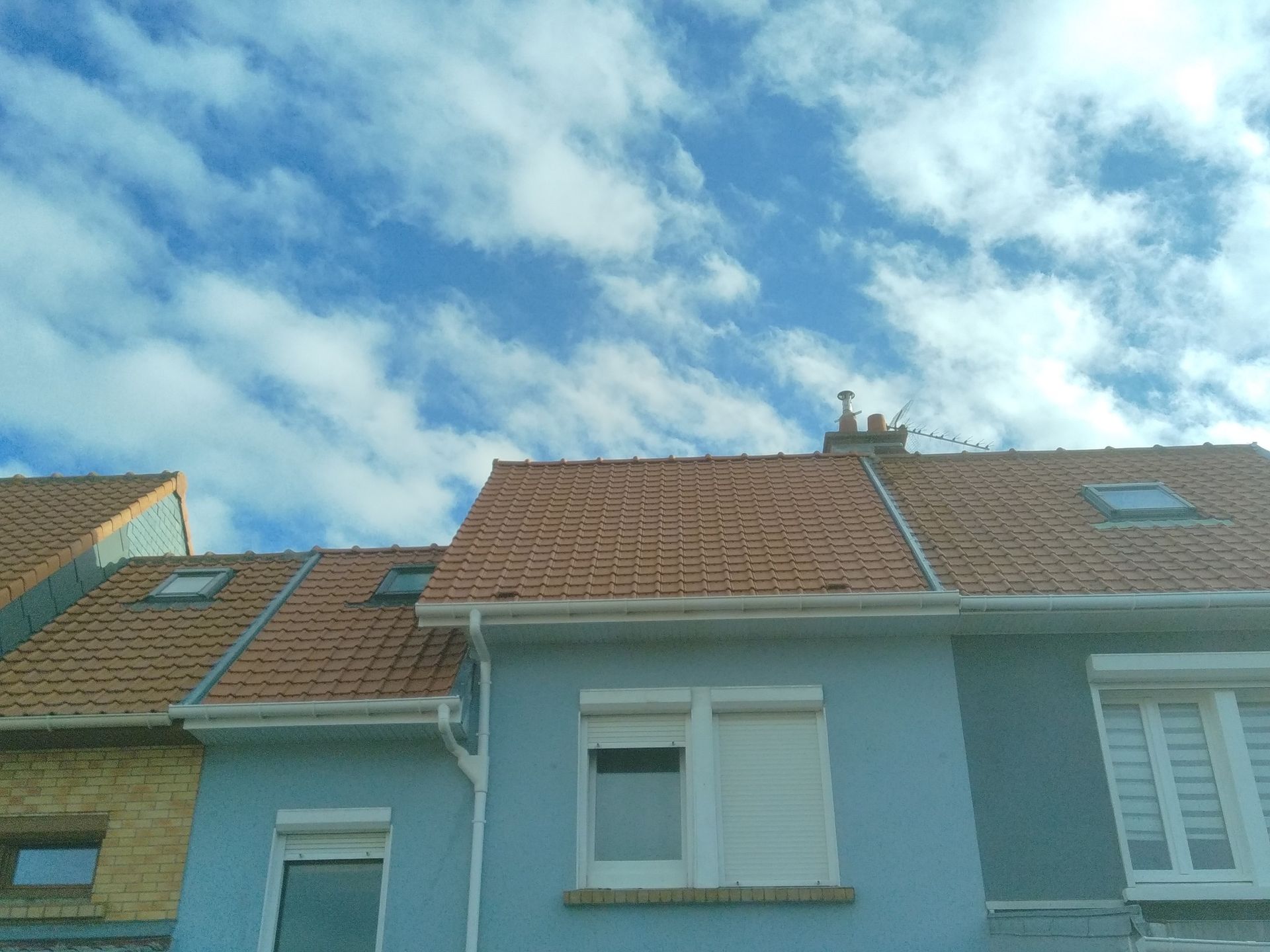 Des maisons bleues aux toits de tuiles orange se détachant sur un ciel bleu parsemé de nuages blancs duveteux.