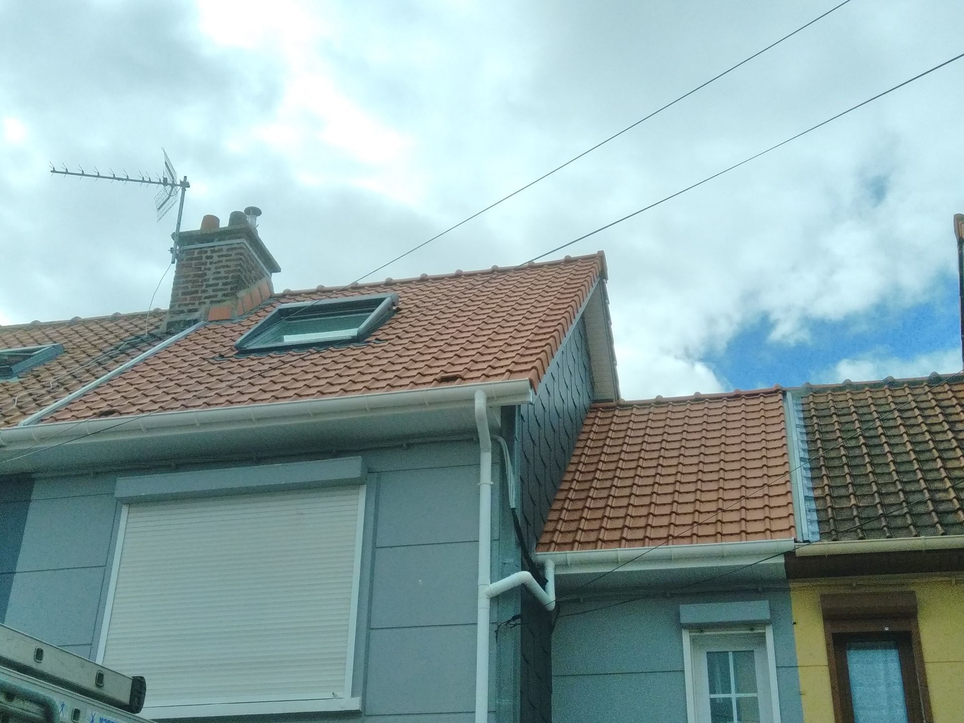 Une maison au toit de tuiles rouges, avec puits de lumière, cheminée et bardage gris, sous un ciel nuageux.