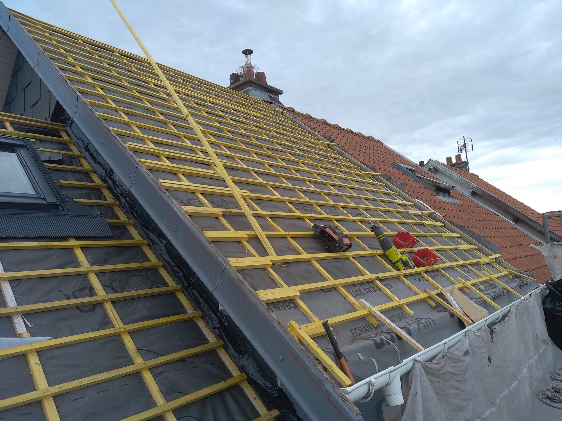 Travaux de toiture en cours : sous-couche noire, charpente jaune, outils, ciel et toit de tuiles rouges.