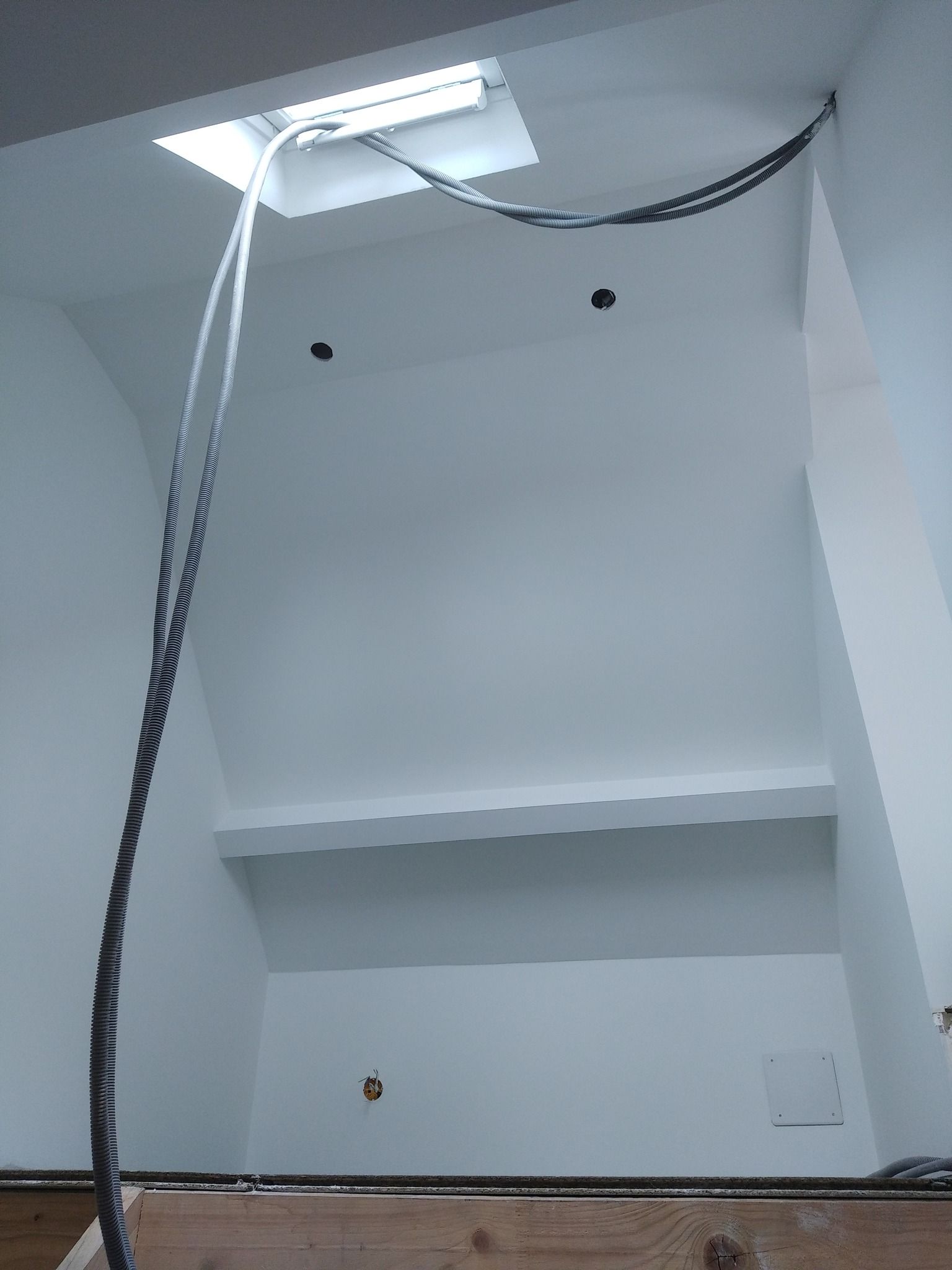 Vue intérieure d'une pièce blanche avec un puits de lumière, des câbles électriques et un escalier en bois partiellement escamoté.