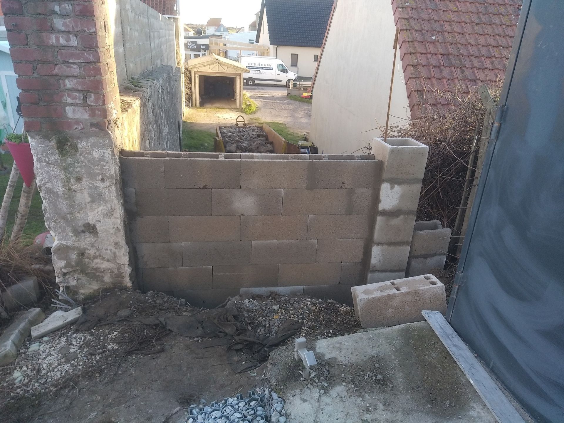 Mur en parpaings en construction, avec des débris à sa base, à côté de structures en briques et d'un quartier résidentiel en arrière-plan.