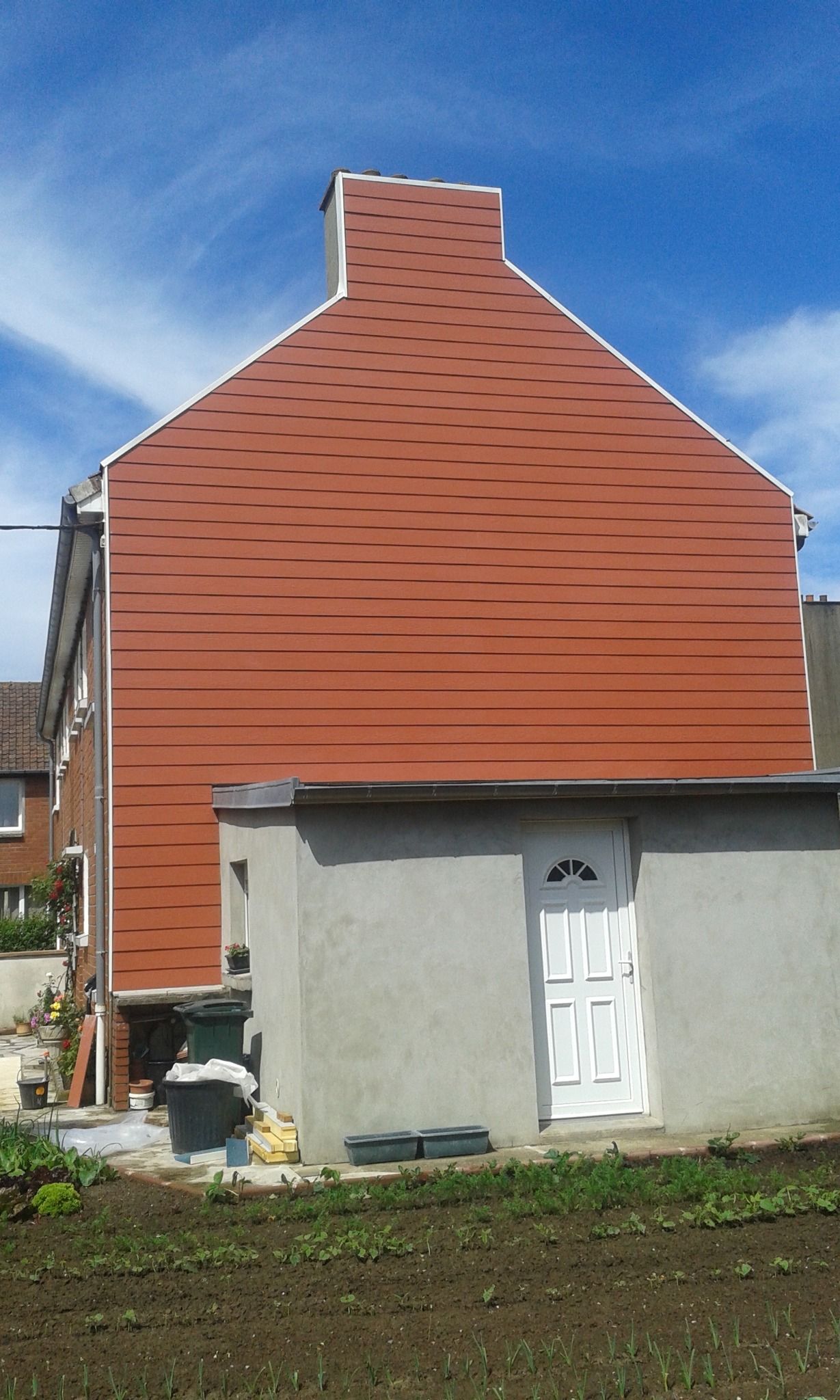 Bâtiment avec un bardage rouge en lignes horizontales, porte blanche et cheminée sur fond de ciel bleu.