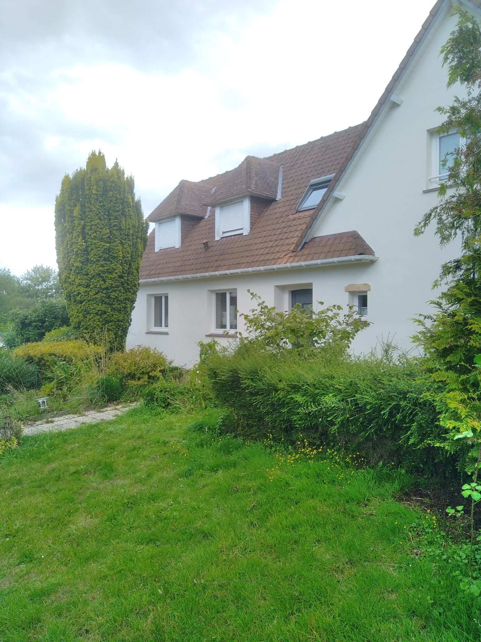 Maison beige avec un toit de tuiles rouges, des fenêtres et un grand arbre vert sur une colline herbeuse.