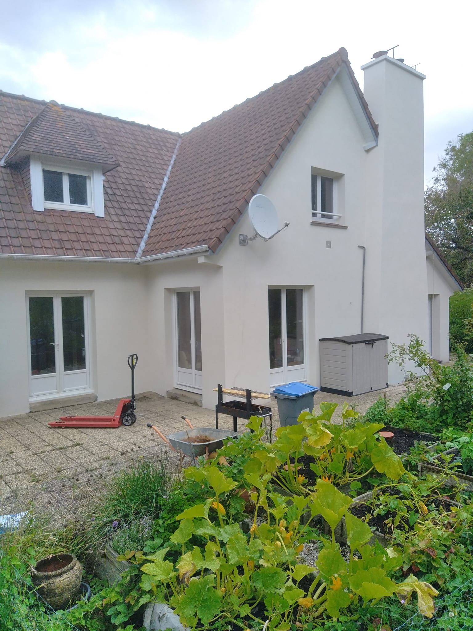 Maison aux murs clairs, au toit de tuiles rouges et à la cheminée. Une antenne parabolique est fixée sur le côté.