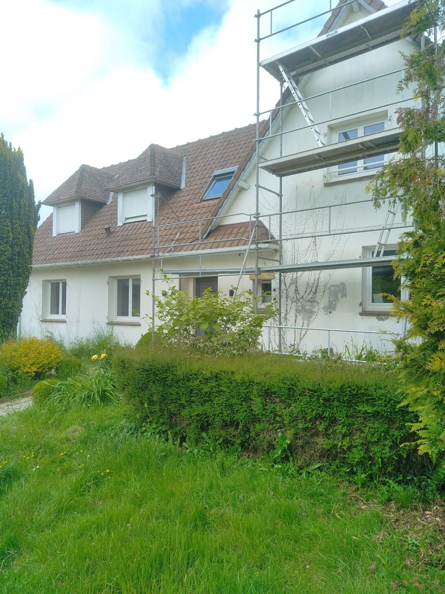 Maison blanche avec échafaudages, toit marron et jardin envahi par la végétation.