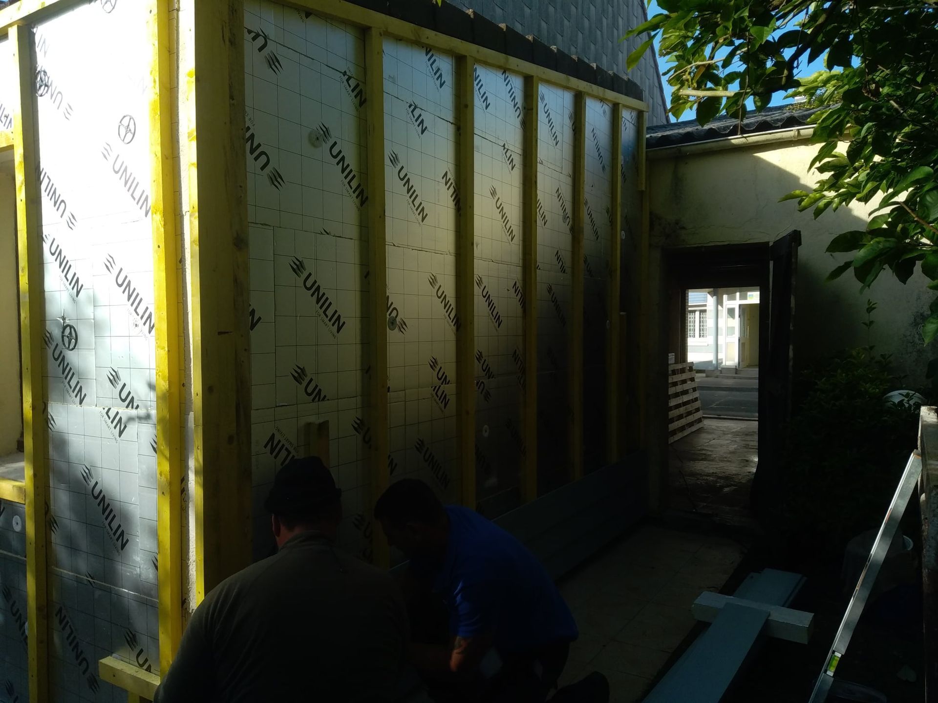 Des ouvriers du bâtiment installent de l'isolant sur un mur extérieur. Charpente en bois jaune, isolant argenté et vue latérale d'un bâtiment.