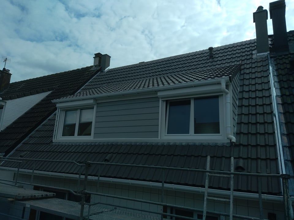 Toiture avec lucarnes, bardage gris et volets sous un ciel nuageux.