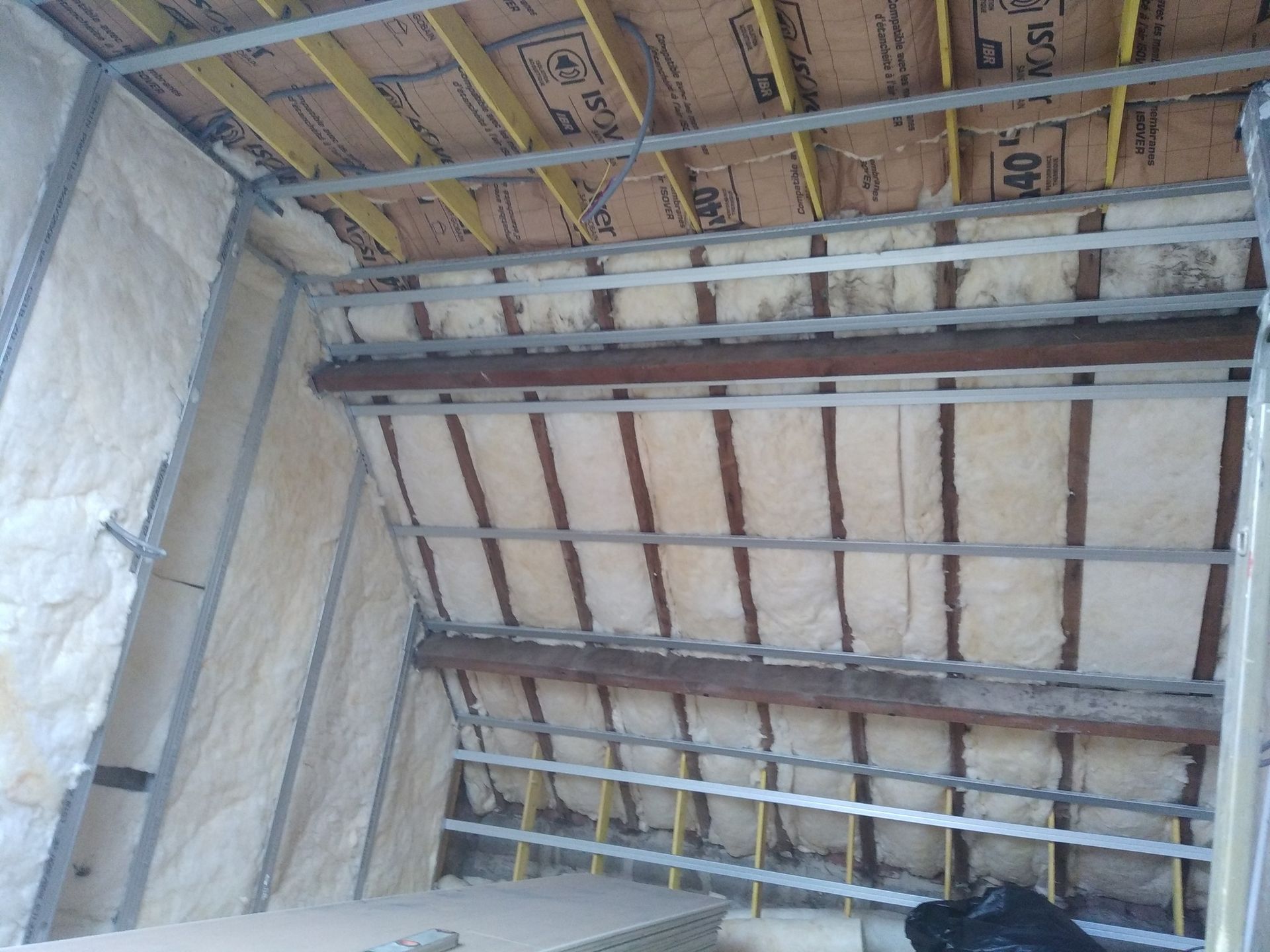 Vue intérieure d'un chantier de construction montrant une ossature métallique, une isolation et des poutres apparentes dans un plafond et un mur en pente.