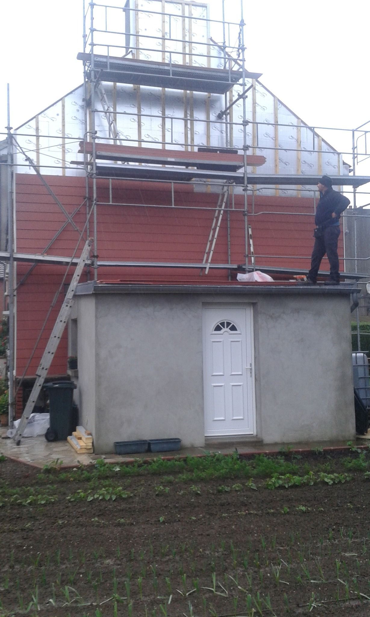 Façade en rénovation avec des bardage rouge et isolation, avec des échafaudages. Une personne se tient sur un petit toit.