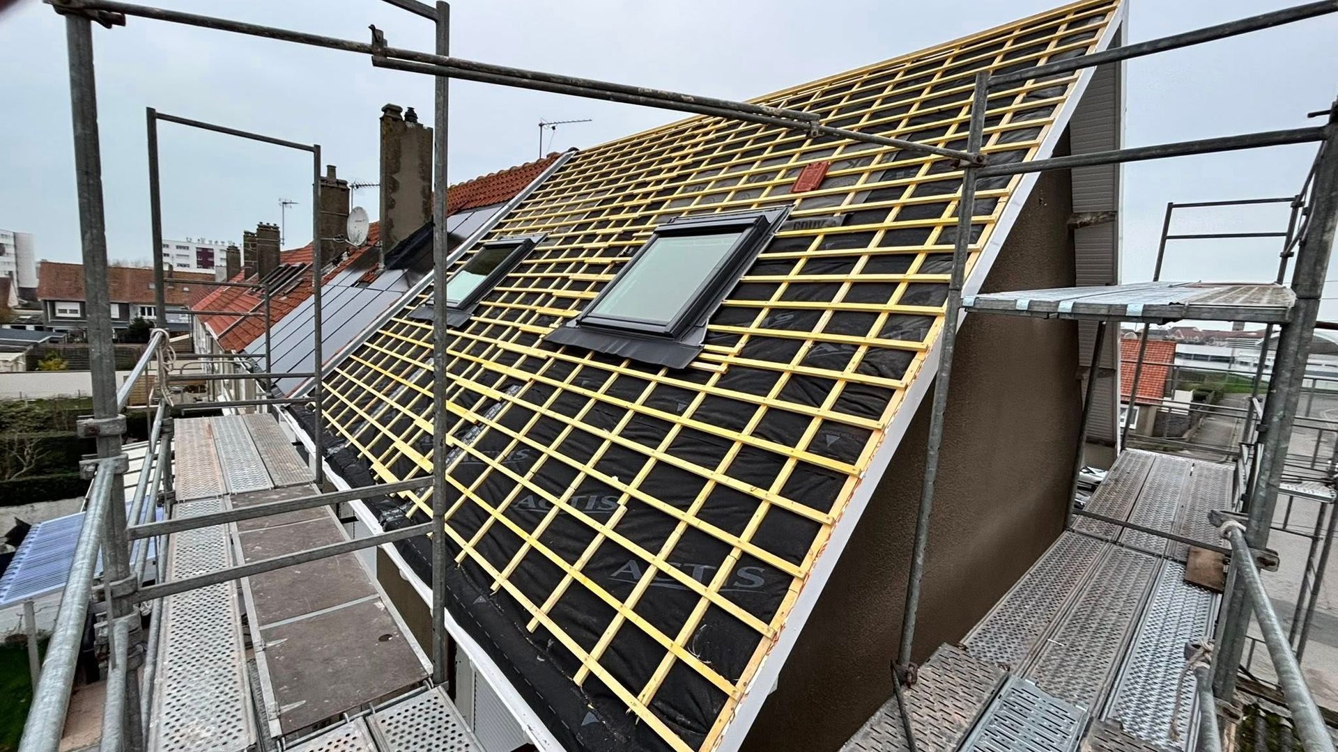 Toiture avec échafaudage, charpente en treillis de bois et deux puits de lumière ; journée nuageuse.