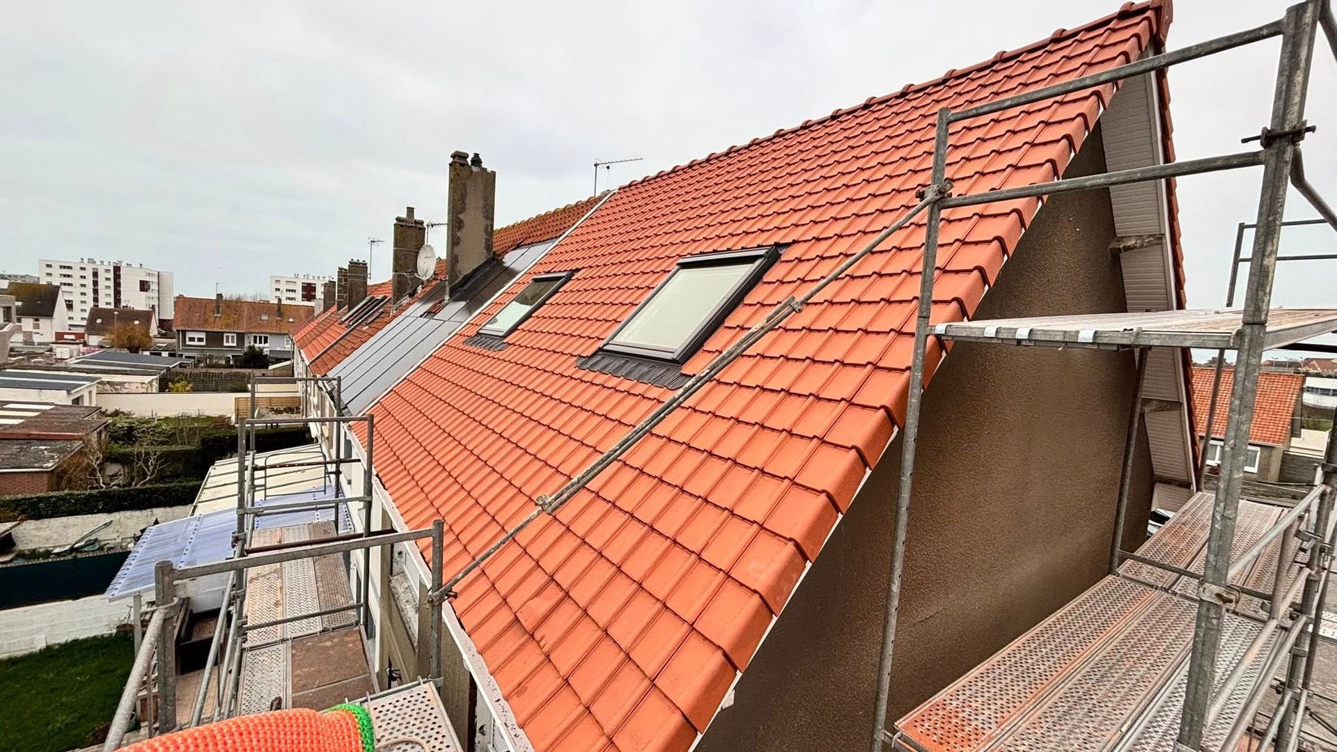 Toit en tuiles rouges avec deux puits de lumière, échafaudages et paysage urbain en arrière-plan.