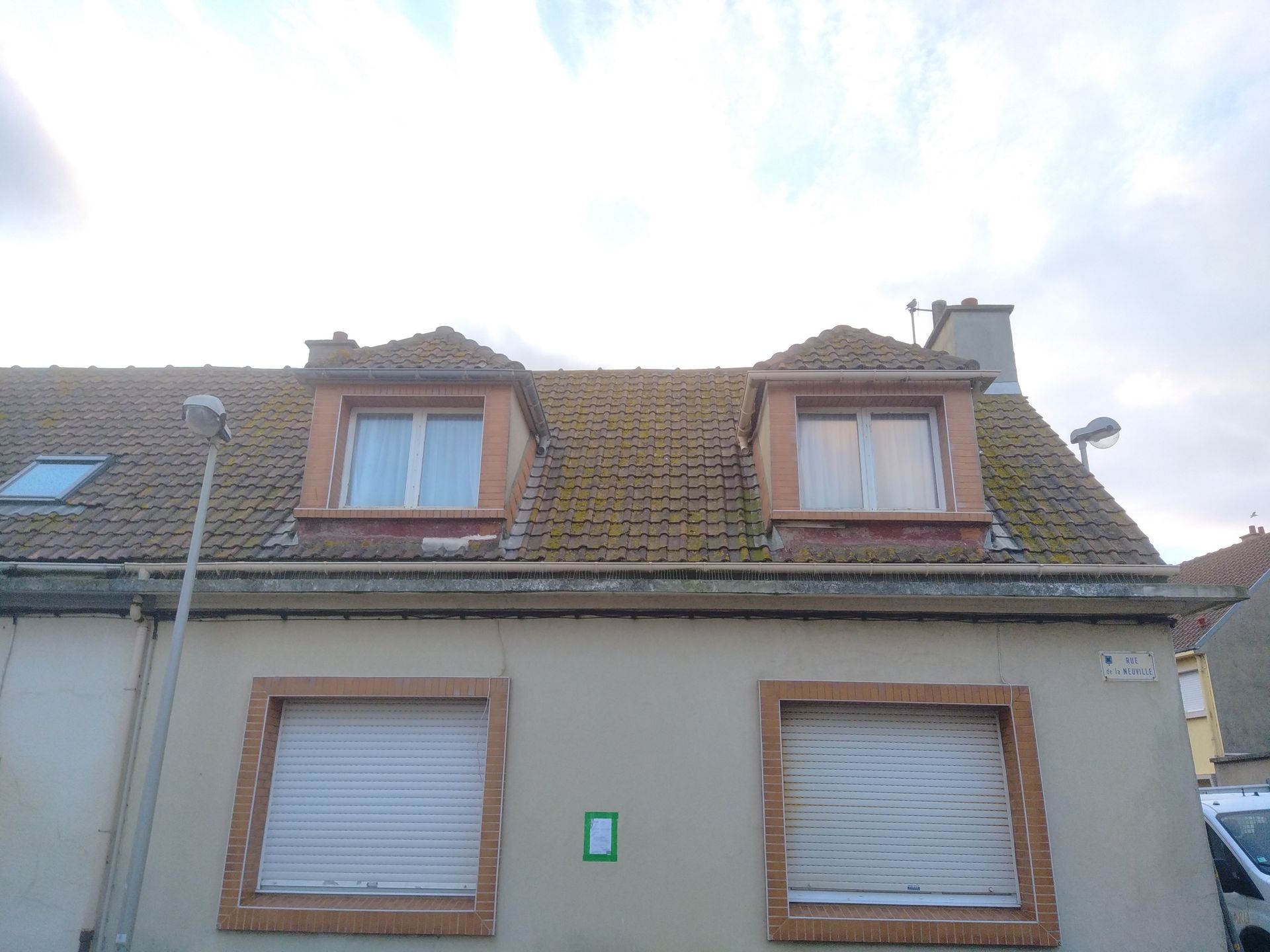 Bâtiment de deux étages avec lucarnes, toit brun et éléments en brique, sous un ciel nuageux.