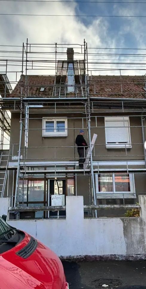 Un homme travaille sur un échafaudage, en façade d'un immeuble. La maison est brune et le ciel est nuageux.