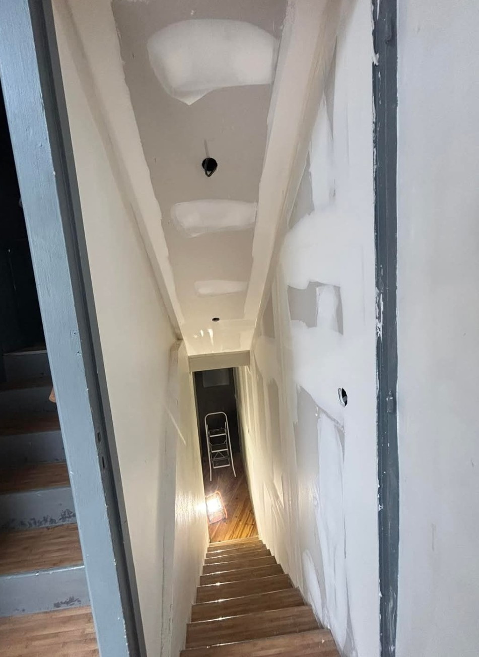 Escalier étroit, murs et plafond blancs, lumière venant du bas, marches visibles.