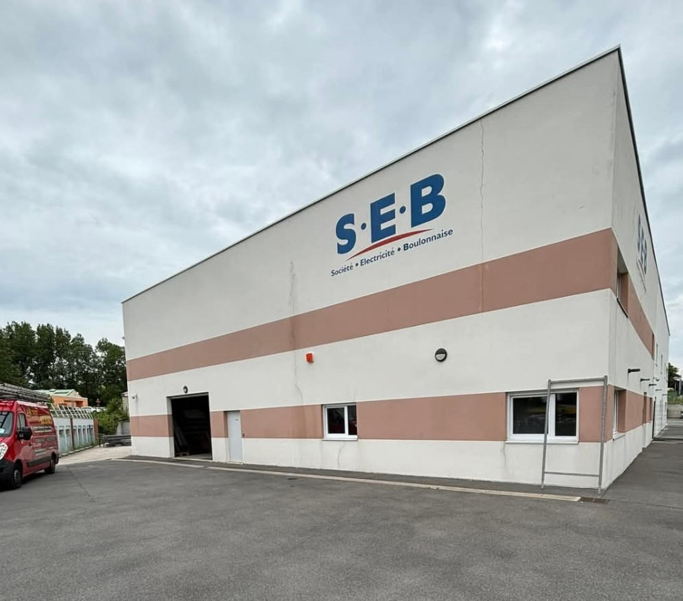 Bâtiment arborant le logo « SEB ». Extérieur blanc et beige, ciel couvert. Camion de pompiers rouge en bas à gauche.