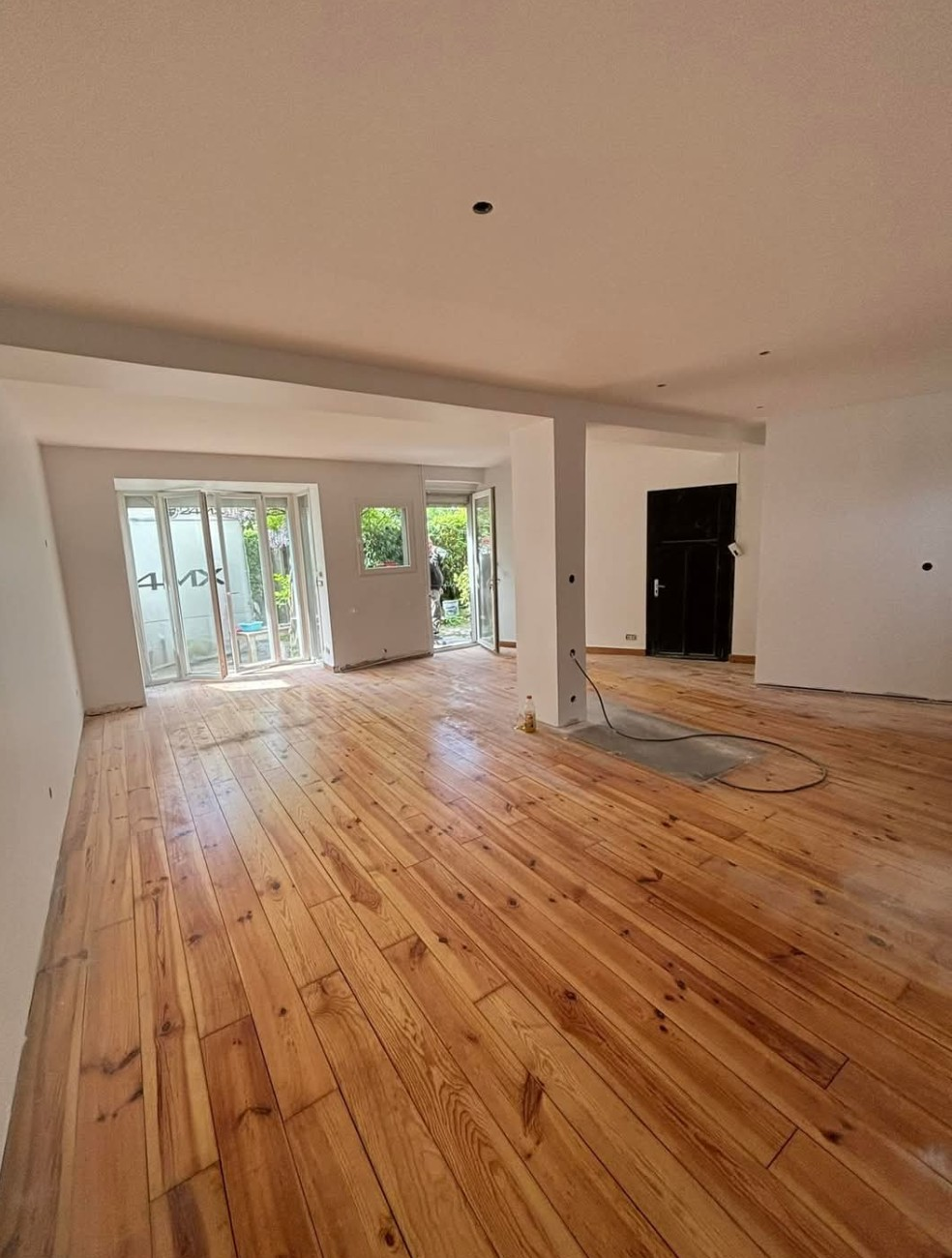 Pièce lumineuse avec parquet récemment rénové et murs blancs, donnant sur un espace extérieur.