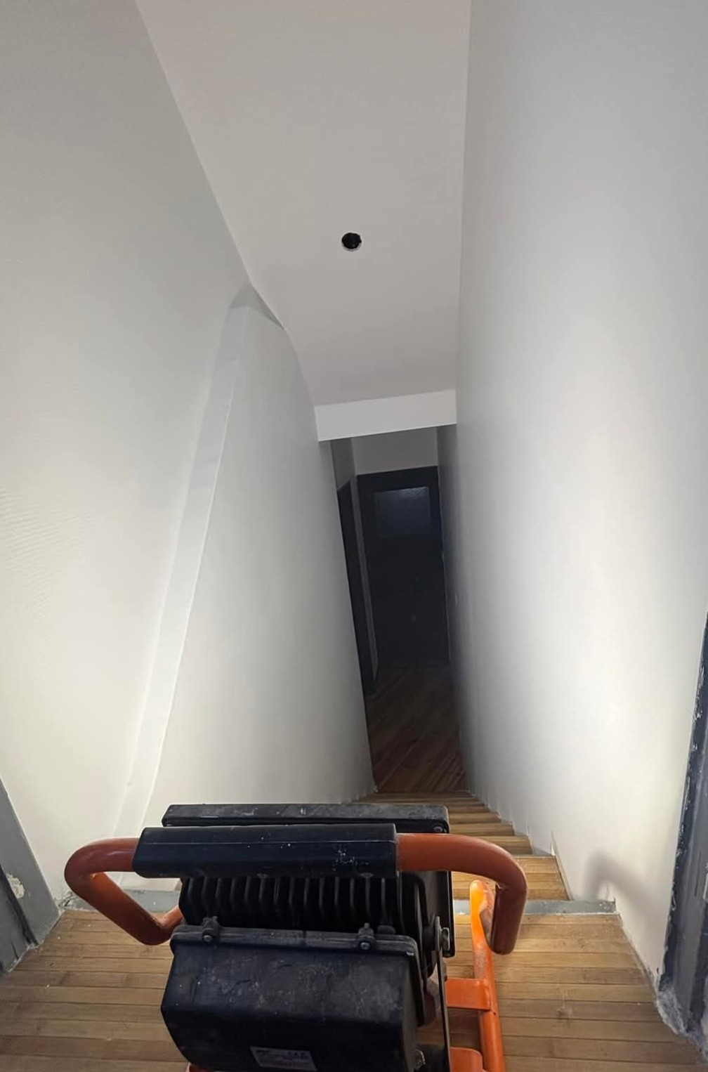 Vue du haut de l'escalier. Un compacteur repose sur des marches en bois, face à une porte sombre.