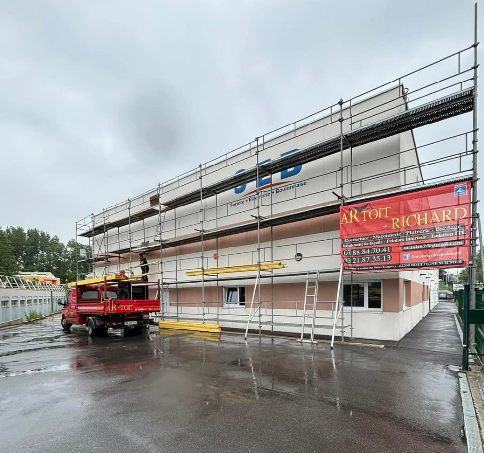 Façade d'un bâtiment en rénovation, avec des échafaudages et un camion rouge garé à proximité.