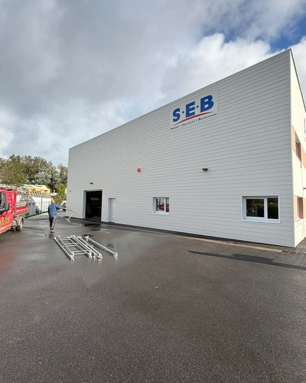 Bâtiment blanc avec l'enseigne « SEB » ; une personne marche près de structures métalliques empilées sur de l'asphalte mouillé.