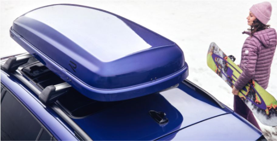 Frau mit Snowboard neben einem blauen Auto mit Dachbox