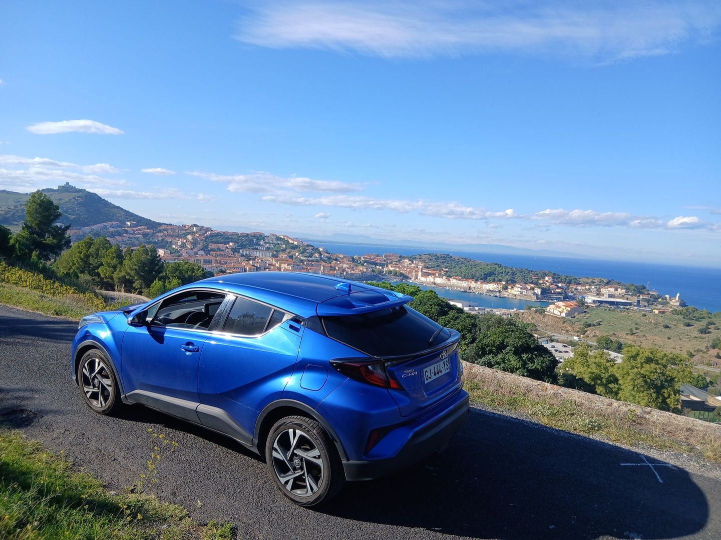 Une Toyota C-HR bleue est garée sur une route surplombant une ville côtière et la mer, sous un ciel d'un bleu éclatant.