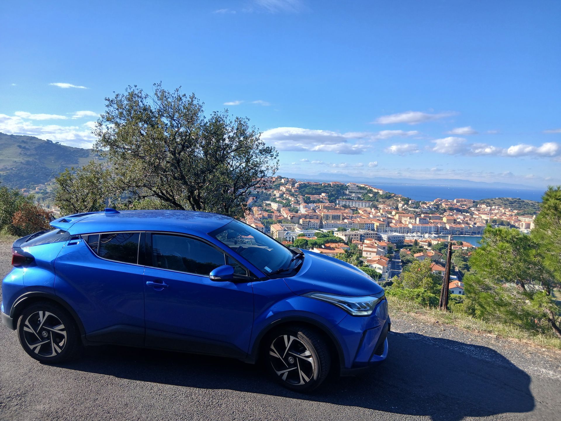 Une Toyota C-HR bleue est garée sur une colline surplombant une ville côtière sous un ciel bleu.