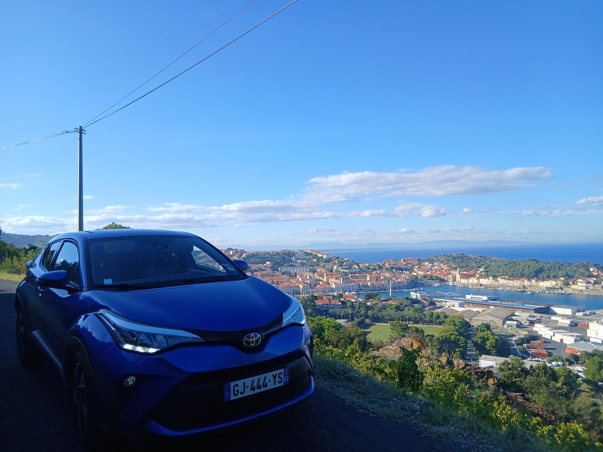 Une Toyota C-HR bleue est garée à flanc de colline, surplombant une ville côtière sous un ciel d'un bleu éclatant.