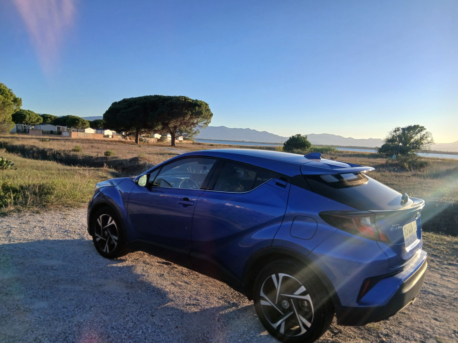 Une Toyota C-HR bleue est garée sur un chemin de terre, des arbres et des montagnes en arrière-plan, sous un ciel bleu.