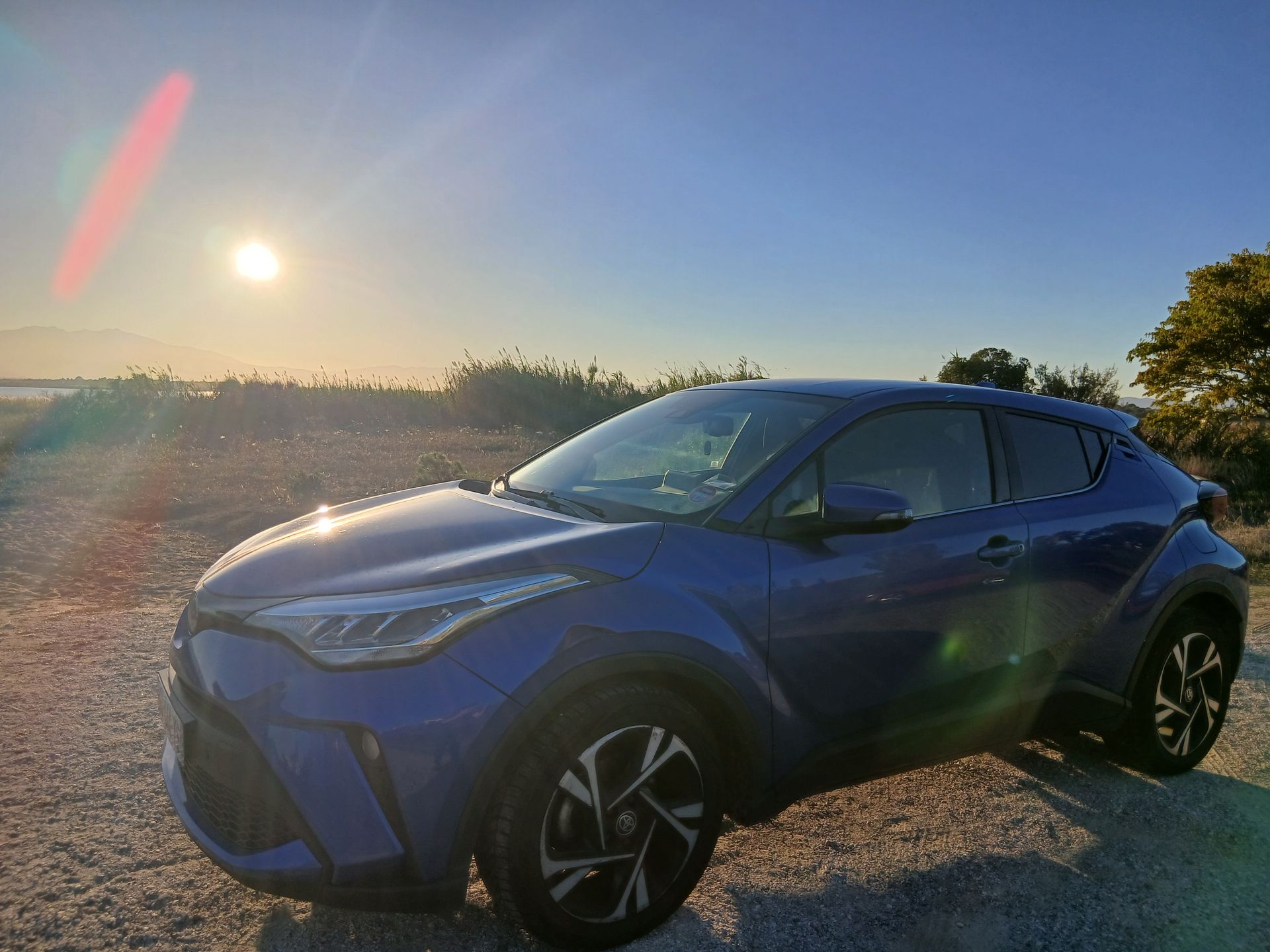 Toyota C-HR bleue garée sur un chemin de gravier, soleil en arrière-plan.
