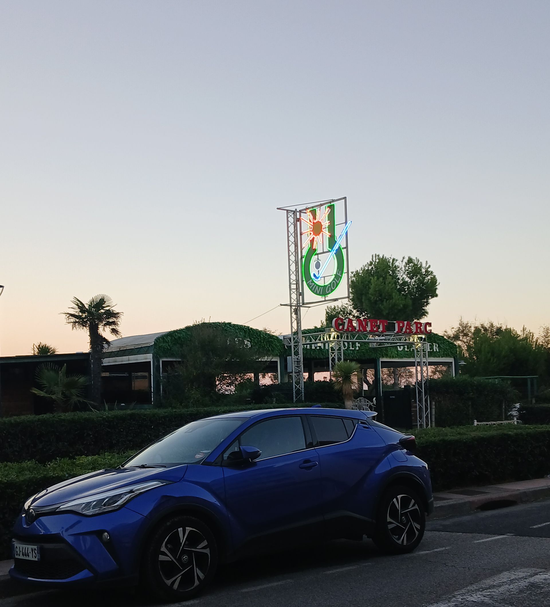 Toyota C-HR bleue garée devant un bâtiment orné d'une grande enseigne. Crépuscule, teintes vertes et beiges.
