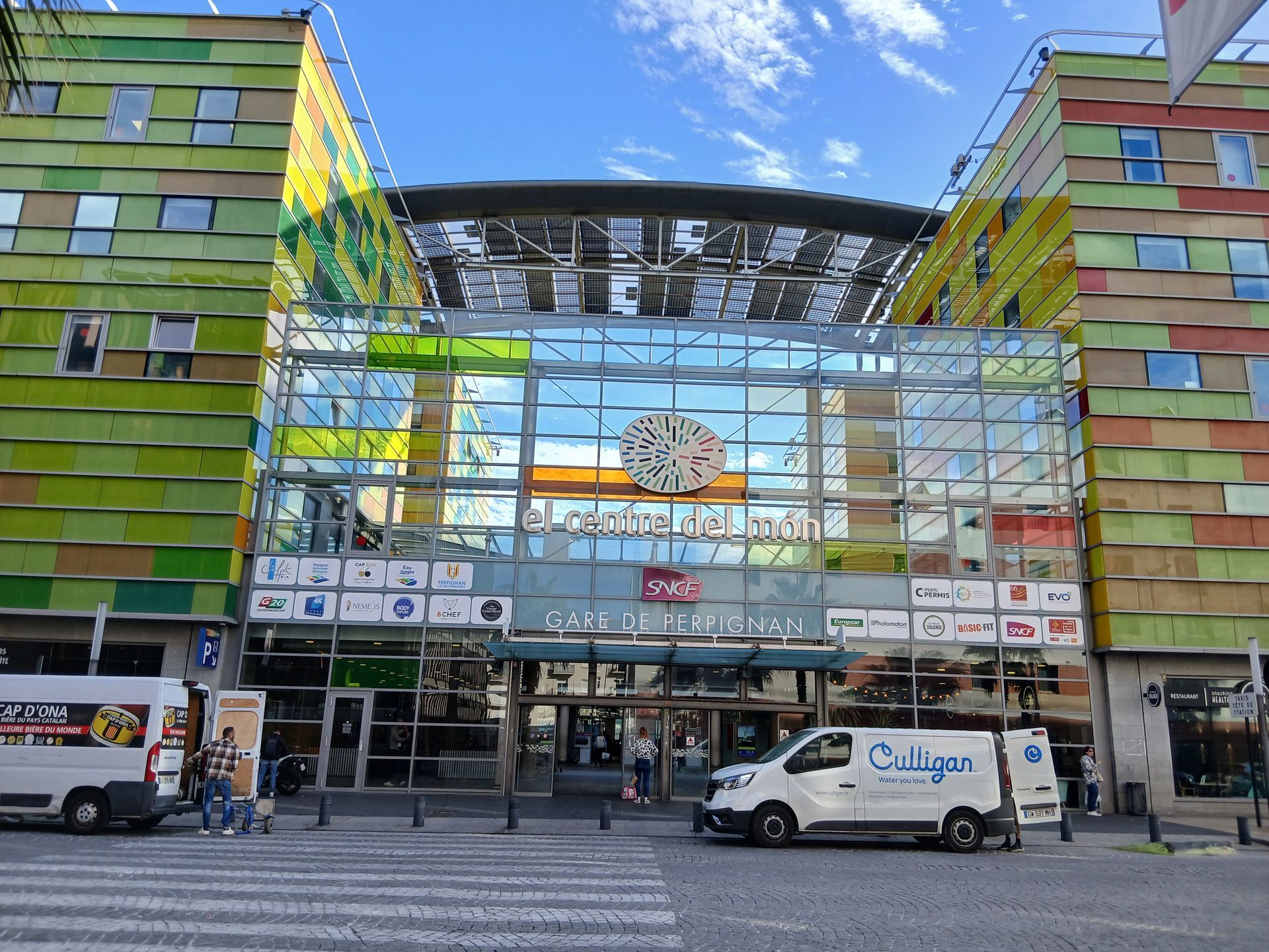 Entrée d'un centre commercial à l'extérieur coloré. Façade vitrée, circulation automobile, ciel bleu.