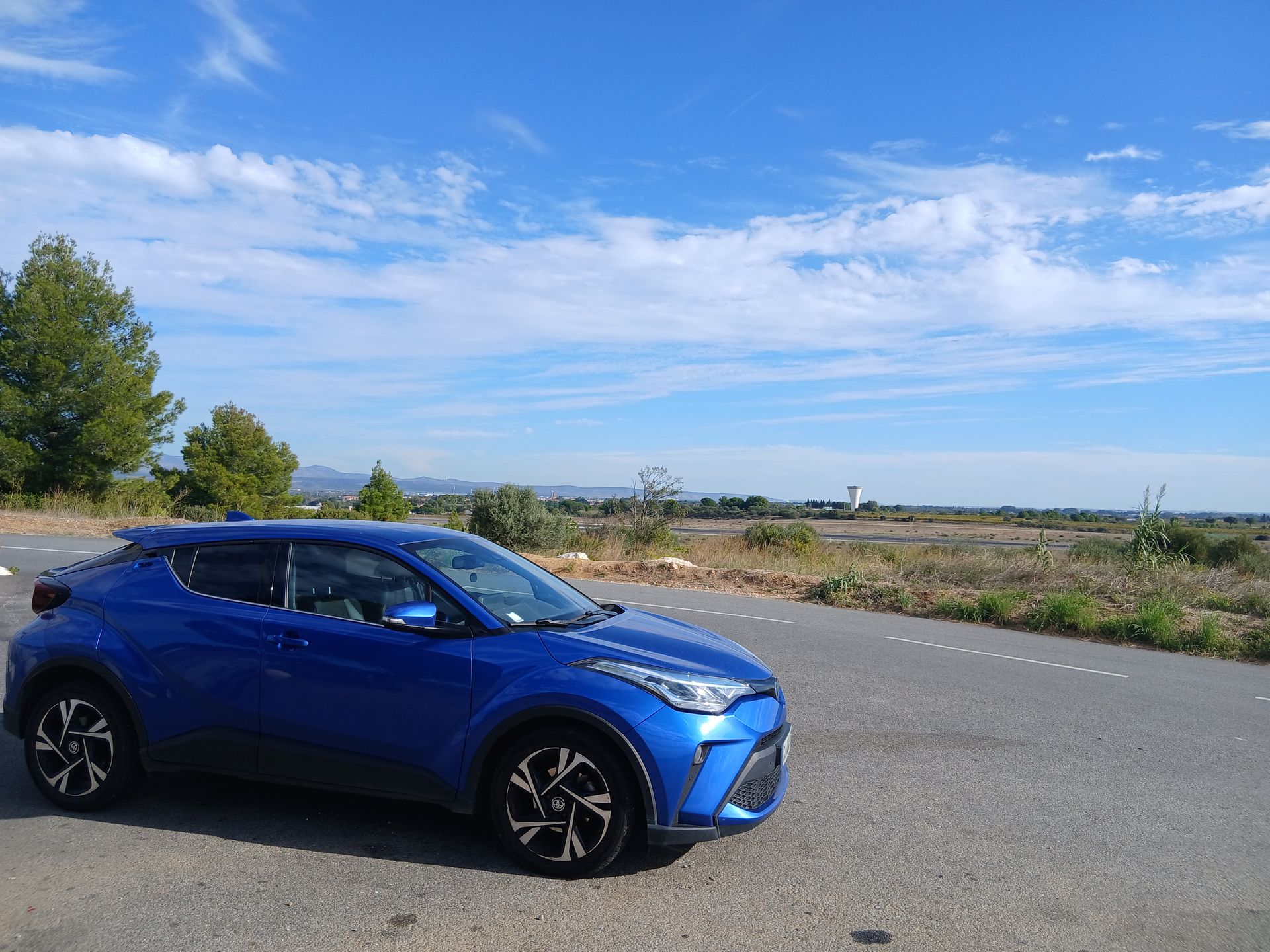 Une Toyota C-HR bleue est garée sur une route, avec un ciel bleu et un paysage au loin.